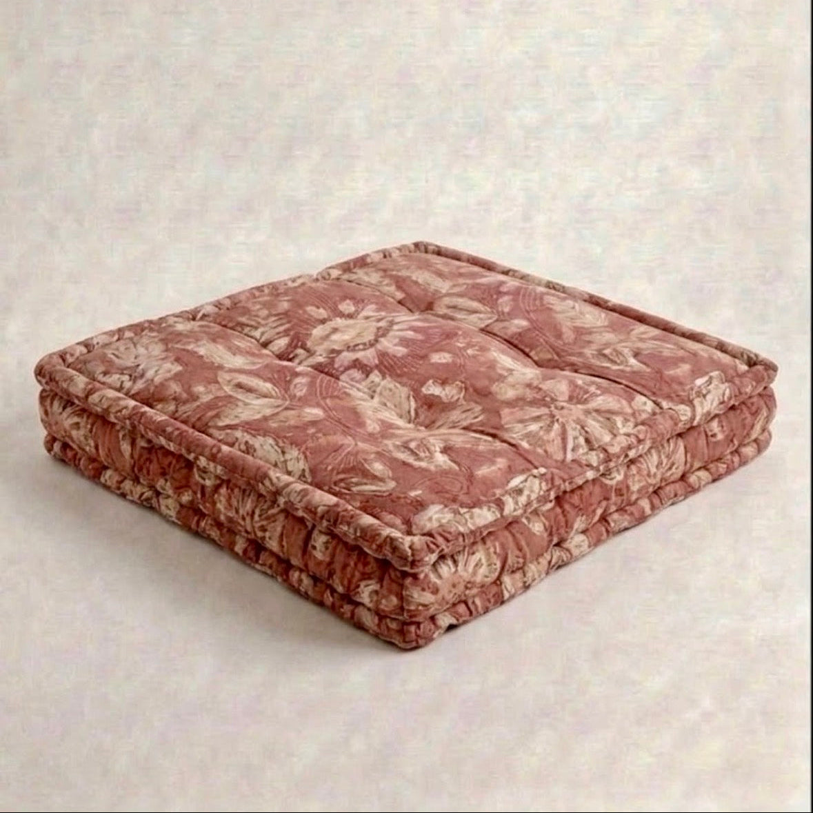 A tufted dog bed in a dusty rose and beige floral pattern, shown on a textured light gray background.
