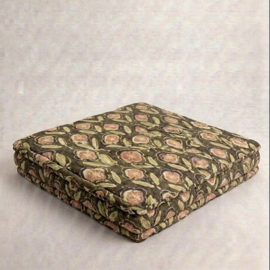 A square upholstered floor cushion with a vintage floral pattern in muted green, pink, and beige tones, shown folded on a textured light gray background.