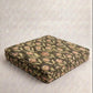 A square upholstered floor cushion with a vintage floral pattern in muted green, pink, and beige tones, shown folded on a textured light gray background.