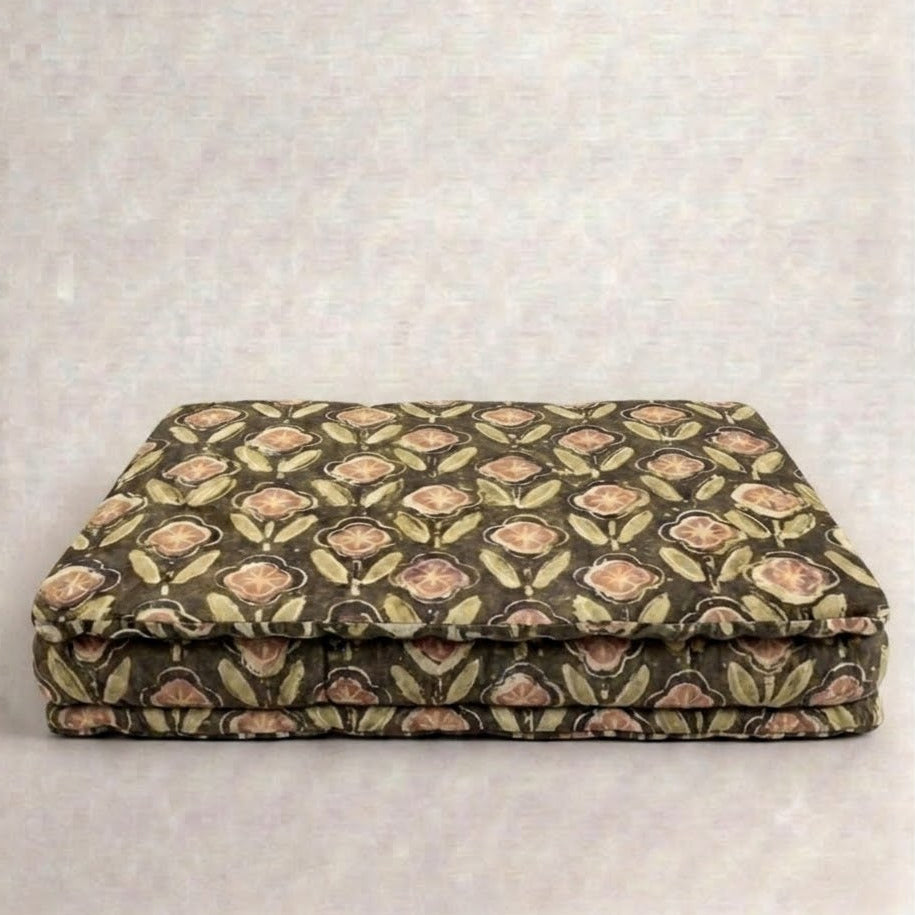A pet bed cushion with a repeating floral pattern in muted green, beige, and brown tones, photographed against a plain light gray textured backdrop.