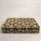 A pet bed cushion with a repeating floral pattern in muted green, beige, and brown tones, photographed against a plain light gray textured backdrop.