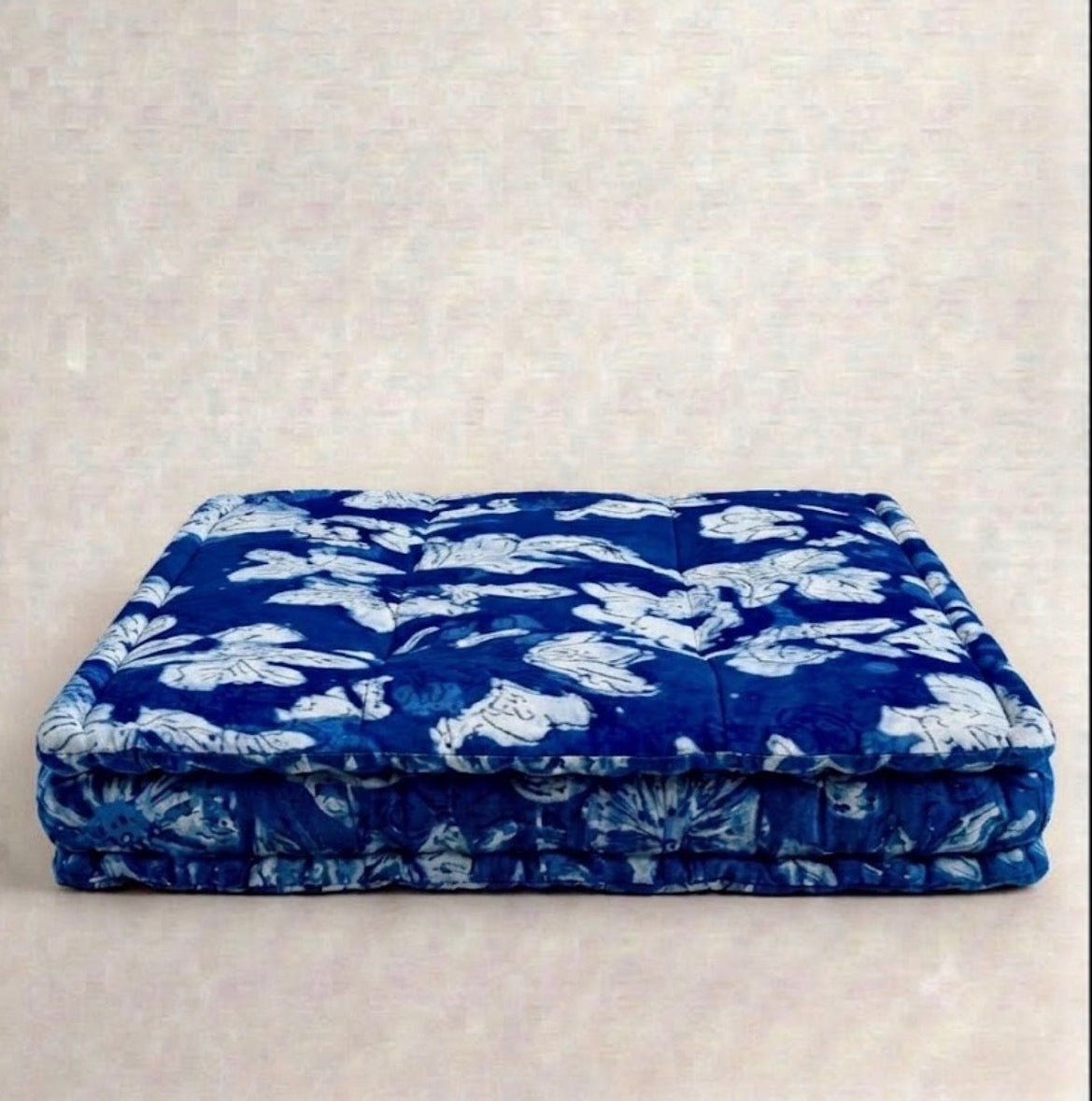 A square blue quilted cushion or floor cushion with a white floral pattern, displayed against a neutral textured beige background.