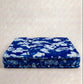 A square blue quilted cushion or floor cushion with a white floral pattern, displayed against a neutral textured beige background.