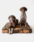 Two dogs sitting on a patterned dog bed against a white background