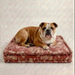 An English Bulldog lying on a square, thick cushioned dog bed with a dusty rose floral pattern, set against a neutral textured beige background.