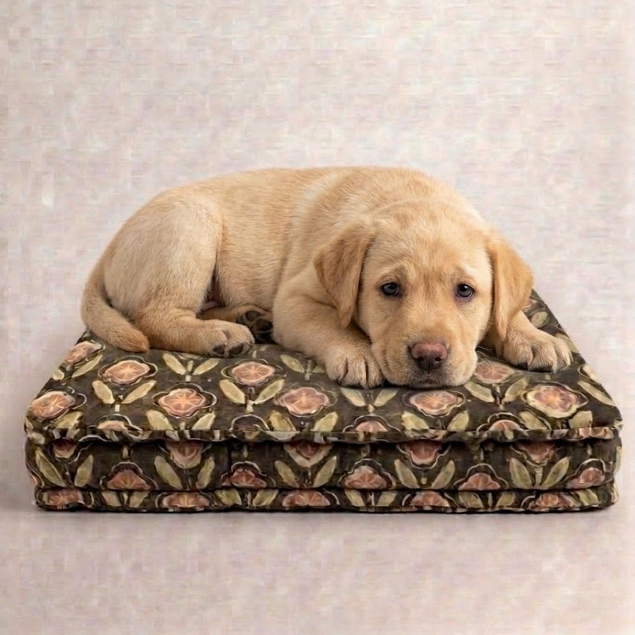 A light cream-colored Labrador puppy resting on a patterned rectangular dog bed with a floral design in brown, beige, and pink tones, set against a neutral textured beige background.