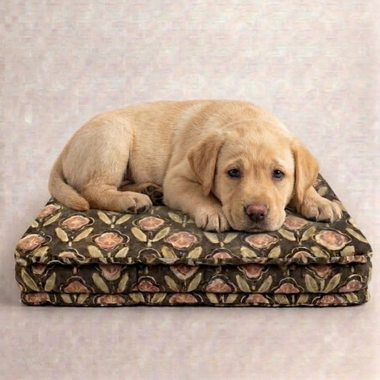 A light cream-colored Labrador puppy resting on a patterned rectangular dog bed with a floral design in brown, beige, and pink tones, set against a neutral textured beige background.
