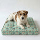 A small brown and white dog lies on a square quilted dog bed with a mint green background and a repeating pattern of small illustrated figures, photographed against a plain light gray studio background.